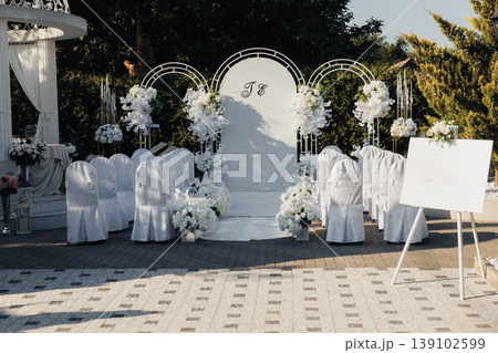 Outdoor Wedding Altar With White Decor And Rows Of Chairs Set On Paved Terrace, Sculptural Backdrop And Abundant Florals Define Ceremony Space Structured Layout, Elegant Staging, Signage Outdoor Wedding Altar With White Decor And Rows Of Chairs Set On Paved Terrace, Sculptural Backdrop And Abundant Florals Define Ceremony Space Structured Layout, Elegant Staging, Signage 139102599