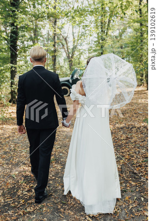 Bride And Groom Walking Hand In Hand Through Woodland, Caucasian Couple Sharing Light Conversation Under Tree Canopy, Umbrella Detail Adding Whimsical Flair And Candid Storytelling Energy. Bride And Groom Walking Hand In Hand Through Woodland, Caucasian Couple Sharing Light Conversation Under Tree Canopy, Umbrella Detail Adding Whimsical Flair And Candid Storytelling Energy. 139102619