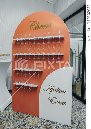 Champagne Glass Display Wall Featuring Rows Of Clear Flutes On Orange Curved Arch, Cheers Header With Apollon Event Script, Elegant Reception Setup For Corporate Launch, Patterned Tile 139102621