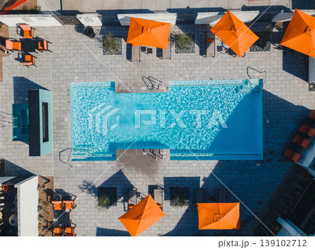 Aerial Rooftop Pool With Orange Umbrellas, Sunlit Turquoise Water, Geometric Tile Deck, Neatly Arranged Lounge Chairs And Parasols, Bold Shadow Patterns, Urban Skyline Glimpses, Curated 139102712
