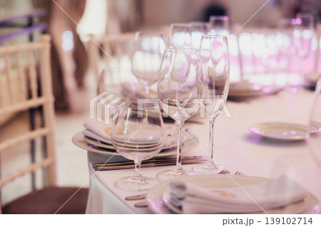 Elegant Table Setting With Glassware, Soft Pink Lighting, Neatly Folded Napkins On White Plates, Crystal Wine Glasses Aligned, Blurred Banquet Chairs In Background, Intimate Romantic Elegant Table Setting With Glassware, Soft Pink Lighting, Neatly Folded Napkins On White Plates, Crystal Wine Glasses Aligned, Blurred Banquet Chairs In Background, Intimate Romantic 139102714