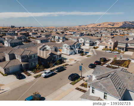 Aerial Suburban Rooftops And Streets Showing Orderly Family Homes, Parked Cars, Manicured Lawns, Solar Panels, Quiet Culdesac, Foothills In Background Under Clear Sky. 139102723