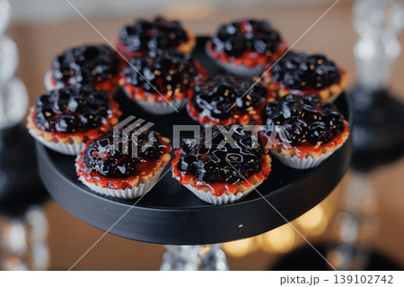 Berry Tarts On Black Stand, Seasonal Brunch Offering With Blueberry Jam Topping And Glossy Glaze, Buttery Crust In Paper Liners, Bitesized Portions For Morning Event, Fresh Fruit Accents, 139102742