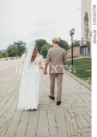 Bride And Groom Walking Along Cobblestone Street, Caucasian Couple Moving Through Historic Urban Setting, Tailored Suit And Flowing Dress, Casual Conversation And Refined Atmosphere. Bride And Groom Walking Along Cobblestone Street, Caucasian Couple Moving Through Historic Urban Setting, Tailored Suit And Flowing Dress, Casual Conversation And Refined Atmosphere. 139102746