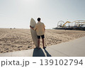 Surfer Leaning On Surfboard Facing Pier, Caucasian Male Acting As Surf Instructor Checking Wave Conditions, Bright Sun, Empty Shoreline, Board Under Arm, Casual Shirt And Shorts, Focused 139102794