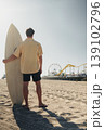 Surfer Standing Near Pier With Surfboard, Caucasian Male Travel Influencer In Casual Beachwear, Textured Sand And Visible Footprints, Vibrant Sky And Ferris Wheel Backdrop, Ready To Paddle 139102796