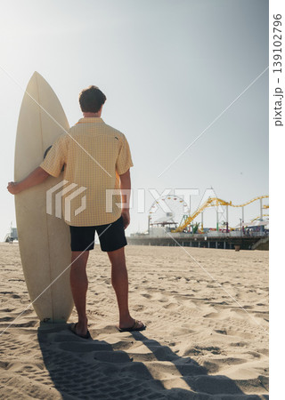 Surfer Standing Near Pier With Surfboard, Caucasian Male Travel Influencer In Casual Beachwear, Textured Sand And Visible Footprints, Vibrant Sky And Ferris Wheel Backdrop, Ready To Paddle Surfer Standing Near Pier With Surfboard, Caucasian Male Travel Influencer In Casual Beachwear, Textured Sand And Visible Footprints, Vibrant Sky And Ferris Wheel Backdrop, Ready To Paddle 139102796