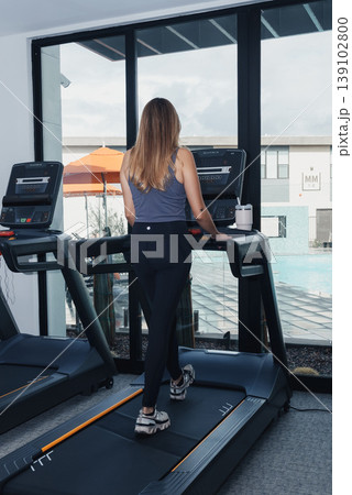Woman Running On Treadmill Overlooking Pool, Sunlit Gym, Relaxed Cardio, Resortstyle Window View, Swim Area Visible, Casual Training Attire, Steady Pace, Cardio Monitoring Display, Wellness 139102800