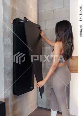 Woman Folding Wallmounted Treadmill Panel, Preparing Home Gym Setup With Careful Adjustment Of Straps, Concrete Wall Backdrop, Beige Activewear, Back View, Compact Storage Solution, Calm 139102801