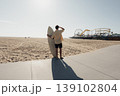 Surfer On Sunlit Boardwalk Holding Surfboard, Caucasian Male Gazing Toward Distant Pier With Long Shadow Across Pavement, Warm Hazy Light, Empty Sand And Calm Ocean, Relaxed Contemplative 139102804
