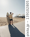 Surfer Casting Long Shadow On Pavement Toward Amusement Pier, Caucasian Male Preparing For Session With Vivid Afternoon Light, Dramatic Silhouette, Distant Ferris Wheel And Empty Sand, 139102805