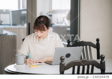 Focused asian woman uses modern technology to work and study sitting at comfortable home table writing notes thoughtfully while looking at laptop screen Focused asian woman uses modern technology to work and study sitting at comfortable home table writing notes thoughtfully while looking at laptop screen 139104476