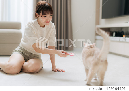 Happy asian woman sitting on living room floor at home taking joyful break from work and study using technology to feed fluffy cat smiling warmly playing with cute domestic pet 139104491