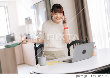 Happy asian woman uses modern technology at home to balance busy work and study routines while smiling talking on mobile phone and cooking food holding pan in bright kitchen 139104496