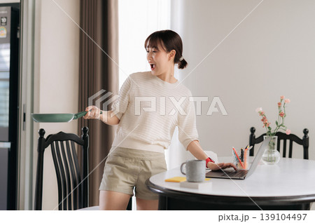 Happy excited asian woman taking break from work and study using technology at home holding green frying pan preparing delicious meal near white table with laptop 139104497