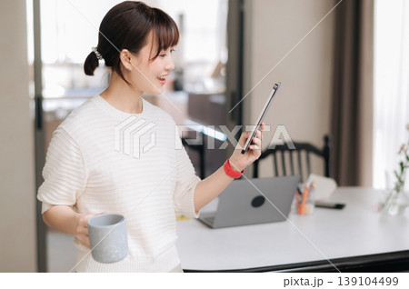 Beautiful happy asian woman uses modern technology for daily work and study at home smiling cheerfully while holding smartphone and coffee mug near laptop computer Beautiful happy asian woman uses modern technology for daily work and study at home smiling cheerfully while holding smartphone and coffee mug near laptop computer 139104499