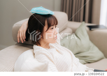 Exhausted asian woman rests on cozy home couch feeling sick with high fever holding ice pack on head after difficult day of remote work and tiring technology study session Exhausted asian woman rests on cozy home couch feeling sick with high fever holding ice pack on head after difficult day of remote work and tiring technology study session 139104522