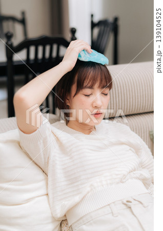 Exhausted asian woman resting at home with painful headache applying cold compress to forehead taking break from technology work and study showing extreme discomfort and fatigue 139104525