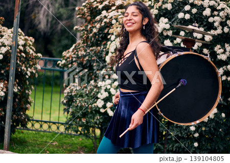 Beautiful dark-haired Latina woman posing with a drum and cymbals on her back, full of joy, a chinchinera, a street musician who dances and plays her drum in the streets. Chilean heritage Beautiful dark-haired Latina woman posing with a drum and cymbals on her back, full of joy, a chinchinera, a street musician who dances and plays her drum in the streets. Chilean heritage 139104805