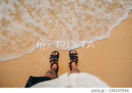 POV looking down at feet in black sandals standing on a sandy beach with ocean waves. POV looking down at feet in black sandals standing on a sandy beach with ocean waves. 139107048