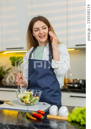 Middle-aged woman on smartphone mixing fresh vegetable salad in kitchen for healthy weight loss diet Middle-aged woman on smartphone mixing fresh vegetable salad in kitchen for healthy weight loss diet 139108114