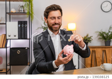 Middle-aged businessman feeds piggy bank with coins dreams travel budget and builds savings plan big Middle-aged businessman feeds piggy bank with coins dreams travel budget and builds savings plan big 139108125