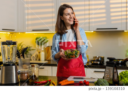 Young woman mixing vegetable salad while talking on smartphone in kitchen at home for healthy diet Young woman mixing vegetable salad while talking on smartphone in kitchen at home for healthy diet 139108310