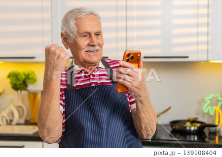 Senior old man celebrating win on smartphone in home kitchen while making healthy fresh diet meal Senior old man celebrating win on smartphone in home kitchen while making healthy fresh diet meal 139108404