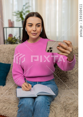 Young woman on sofa wearing headphones watching video lesson on mobile phone taking study notes Young woman on sofa wearing headphones watching video lesson on mobile phone taking study notes 139108411