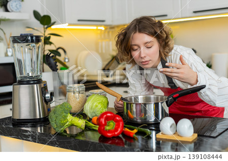 Young woman smelling healthy vegetable soup in home kitchen while cooking dinner for balanced diet Young woman smelling healthy vegetable soup in home kitchen while cooking dinner for balanced diet 139108444