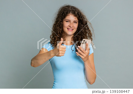 Woman scrolling smartphone news feed smiling giving thumbs up like gesture positive review reaction Woman scrolling smartphone news feed smiling giving thumbs up like gesture positive review reaction 139108447