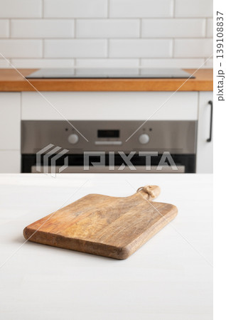 Wooden cutting board on light kitchen table with white kitchen and oven in the background. 139108837