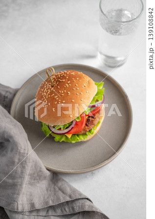 Beef burger with tomato, cheese, and lettuce on a plate on a kitchen table with glass of water Beef burger with tomato, cheese, and lettuce on a plate on a kitchen table with glass of water 139108842