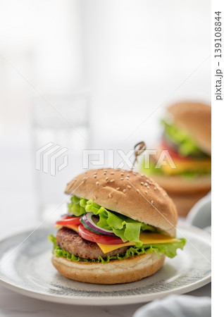 Two Beef burgers with tomato, cheese, and lettuce on a plate on a table near window  139108844