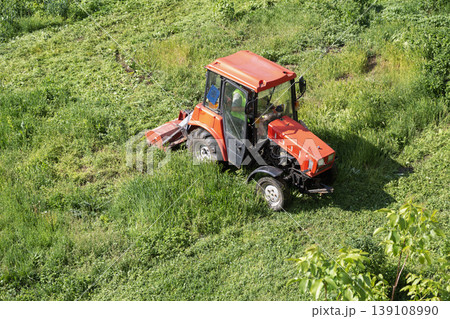 A small red tractor with a lawnmower cutting grass on a lawn. A small red tractor with a lawnmower cutting grass on a lawn. 139108990