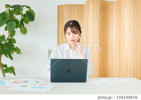 オフィスでノートパソコンを見ながら考える若い女性ビジネスパーソン・資料とグラフを広げたデスクワーク風 オフィスでノートパソコンを見ながら考える若い女性ビジネスパーソン・資料とグラフを広げたデスクワーク風 139109656