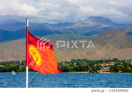 National flag of Kyrgyzstan on pole by serene Issyk-Kul lake with mountains and village 139109992
