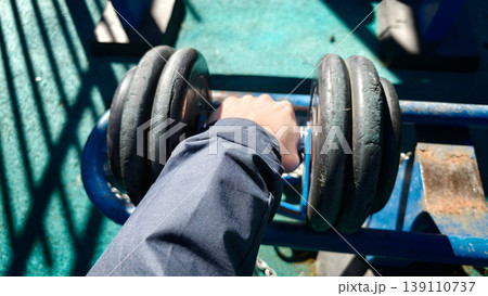 Person lifts dumbbell on outdoor gym equipment in bright daylight 139110737