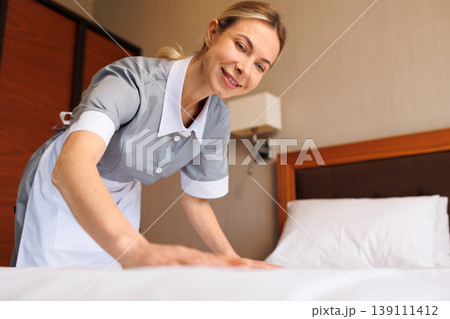 Close up of smiling hotel housekeeper making neatly arranged bed Close up of smiling hotel housekeeper making neatly arranged bed 139111412