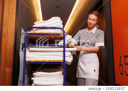 Professional hotel housekeeper arranging clean linens on service cart in modern corridor near guest room door Professional hotel housekeeper arranging clean linens on service cart in modern corridor near guest room door 139111637
