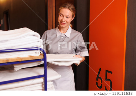 Smiling hotel maid carrying fresh bed sheets from housekeeping trolley beside room number sign 139111638