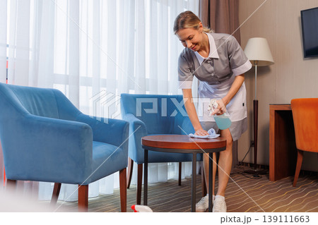 Professional female cleaner wiping round coffee table in modern hotel suite 139111663