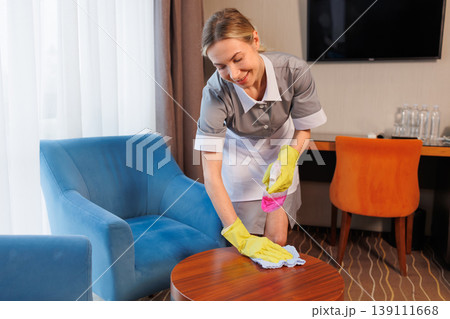 Professional maid disinfecting coffee table in stylish hotel room with blue armchair and modern interior Professional maid disinfecting coffee table in stylish hotel room with blue armchair and modern interior 139111668