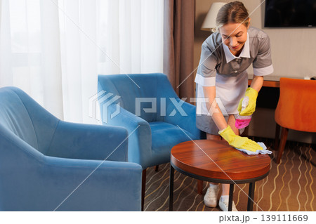 Female cleaner wiping wooden coffee table with disinfectant spray in elegant hotel lounge room 139111669
