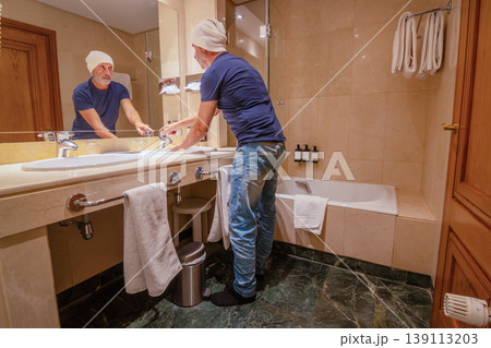 A bearded man wearing a blue t-shirt and white beanie washes his hands at a marble vanity in a luxury hotel bathroom A bearded man wearing a blue t-shirt and white beanie washes his hands at a marble vanity in a luxury hotel bathroom 139113203