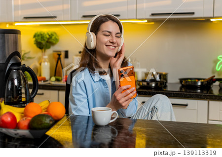 Young woman listening to music with headphones using smartphone in home kitchen relaxing with coffee Young woman listening to music with headphones using smartphone in home kitchen relaxing with coffee 139113319