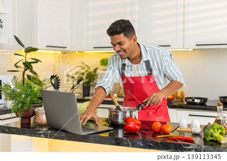 Indian man making healthy soup while looking at laptop in home kitchen for weight loss meal routine 139113345