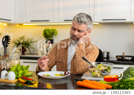 Middle-aged man grimacing while eating healthy salad forcing diet willpower in modern home kitchen 139113365
