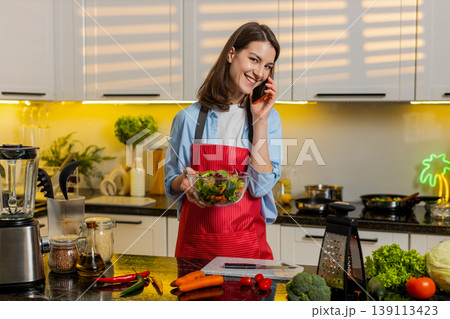 Young woman mixing vegetable salad while talking on smartphone in kitchen at home for healthy diet 139113423