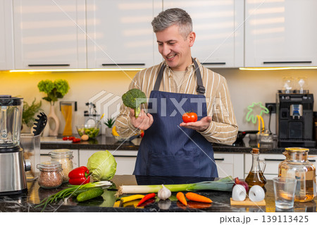 Middle-aged man comparing broccoli and tomato in kitchen while choosing vegetables for healthy meal 139113425
